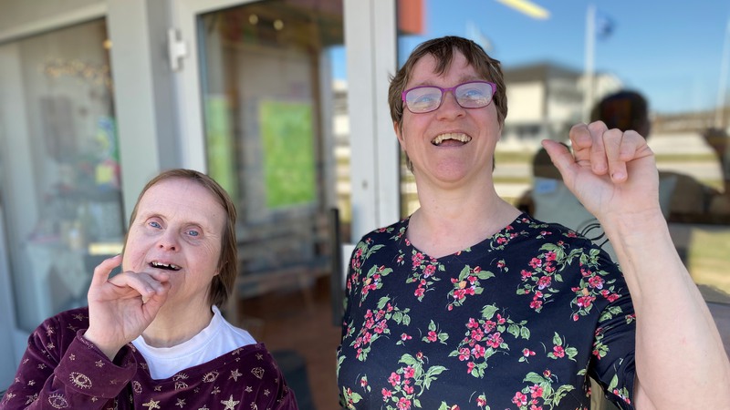 Två glada kvinnor med funktionshinder tittar glatt in i kameran och vinkar