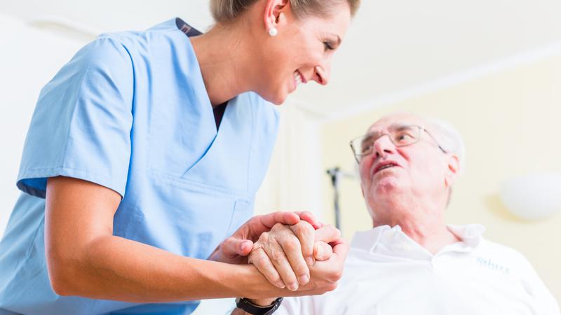 En kvinna som arbetar inom vård eller omsorg håller en äldre mans hand, de tittar på varandra och ler.